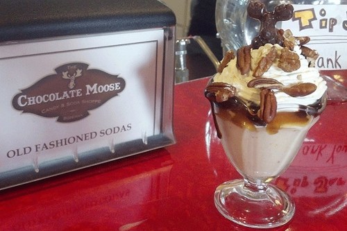 A delicious sundae topped with whipped cream, pecans, and chocolate drizzle, next to a sign for The Chocolate Moose.