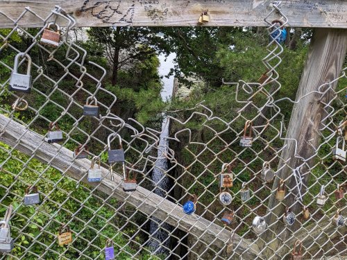 A wooden fence with various padlocks attached, overlooking a small waterfall and lush greenery.