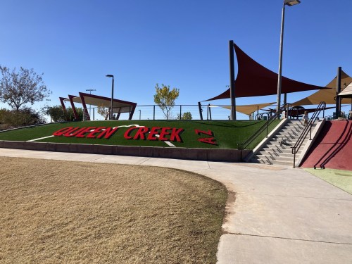 A park in Queen Creek, AZ, featuring green grass, red signage, and shaded play areas under a clear blue sky.