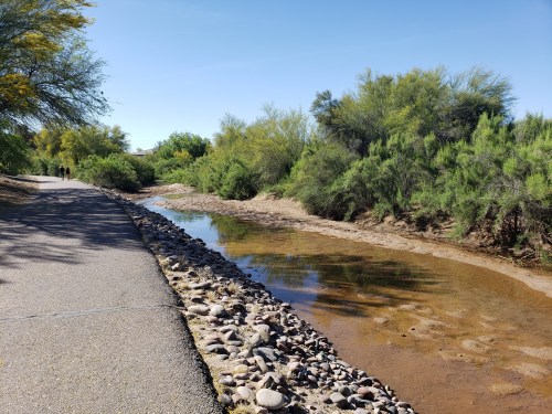 A peaceful pathway alongside a shallow creek, surrounded by lush greenery and clear blue skies.