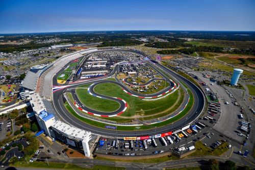 Aerial view of a large racetrack surrounded by parking and facilities, with a clear blue sky above.
