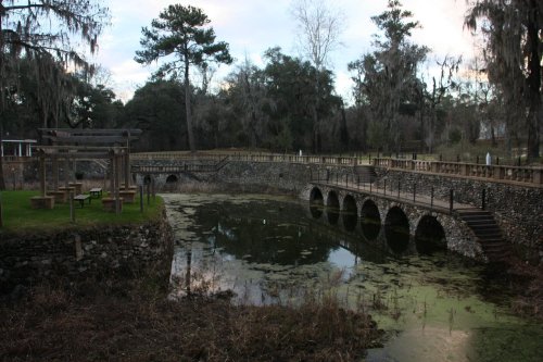 A serene landscape featuring a stone bridge over a pond, surrounded by trees and moss, with a wooden pergola nearby.