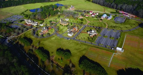Aerial view of a large, green landscape featuring buildings, parking lots, and a pond surrounded by trees.