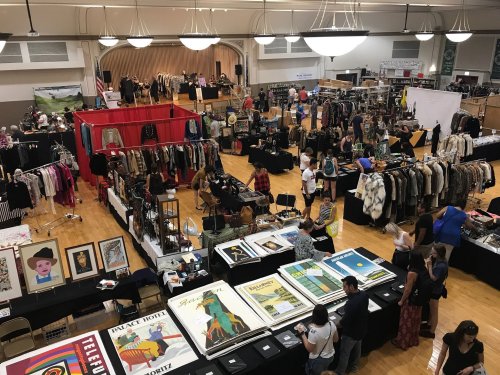 A bustling indoor market with vendors selling clothing, art, and accessories, and shoppers browsing various stalls.