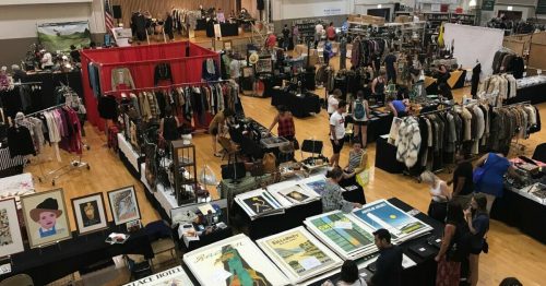 A bustling indoor market with vendors selling clothing, art, and accessories, and shoppers browsing various stalls.