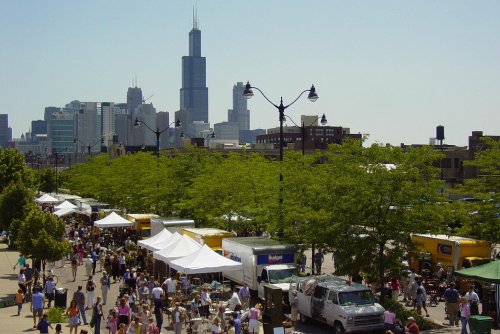 A bustling outdoor market with food trucks and tents, set against a city skyline featuring a tall skyscraper.
