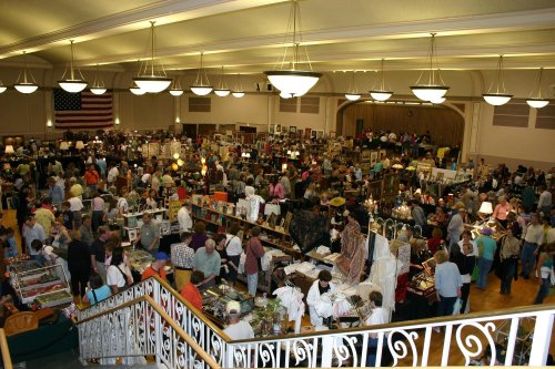 A bustling indoor market with numerous vendors and shoppers browsing various items, under bright lights and an American flag.