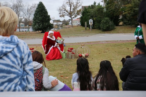 A festive scene with Santa sitting on a chair, surrounded by gifts, while children watch from a distance.
