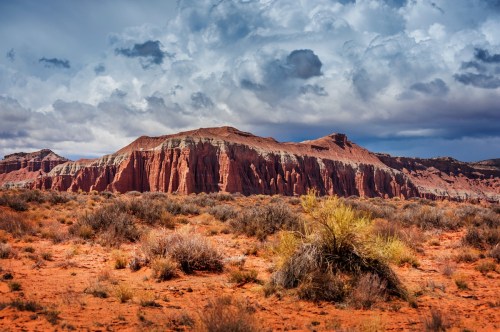 A dramatic landscape featuring red rock formations under a cloudy sky, with sparse vegetation in the foreground.