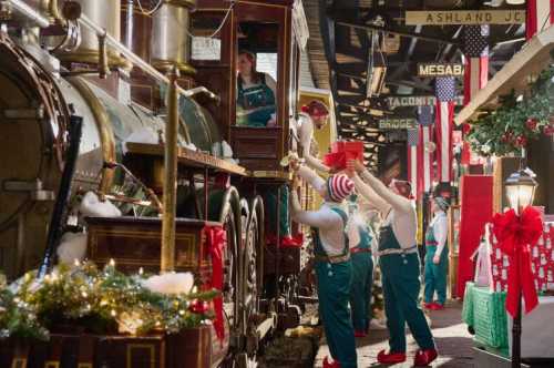 Elves in festive attire load gifts onto a vintage train in a decorated holiday setting.