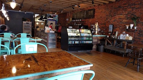 Cozy café interior with brick walls, wooden tables, and a display case filled with drinks. Customers are seated and ordering.