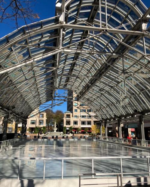 A spacious ice skating rink under a glass-roofed structure, surrounded by buildings and trees on a sunny day.