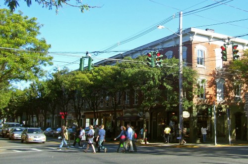 A busy street scene with pedestrians crossing, green traffic lights, and brick buildings under a clear blue sky.