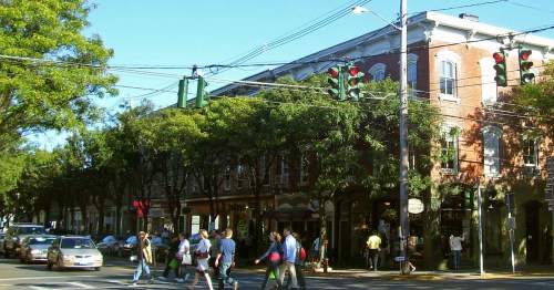 A busy street scene with pedestrians crossing, green traffic lights, and brick buildings under a clear blue sky.
