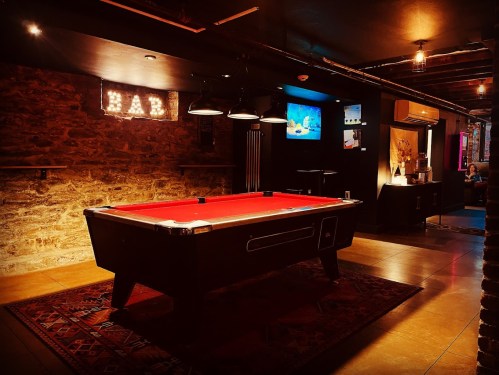 A dimly lit bar area featuring a red pool table, stone walls, and a "BAR" sign in the background.