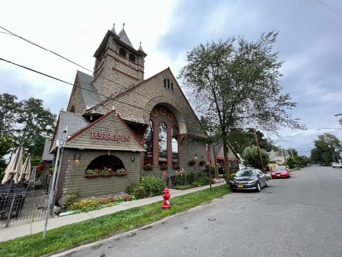 A charming restaurant with a unique architectural design, surrounded by greenery and parked cars on a quiet street.