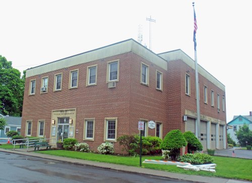 Brick building with a flagpole, featuring large windows and a sign for the municipal office, surrounded by greenery.