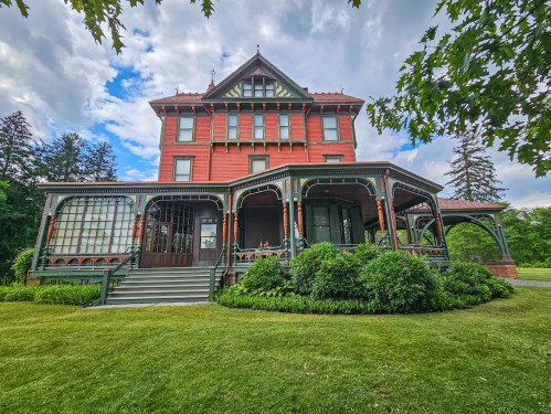 A large red Victorian-style house with intricate woodwork, surrounded by lush greenery and a blue sky.