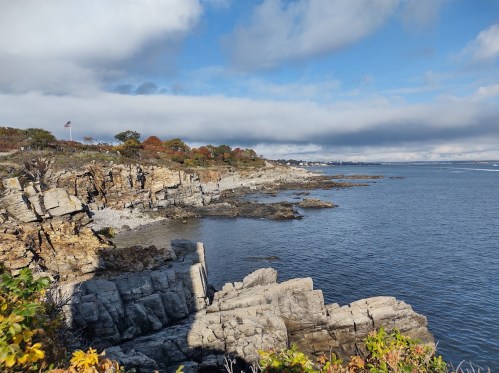 Coastal landscape with rocky cliffs, calm water, and a cloudy sky, featuring autumn foliage along the shore.