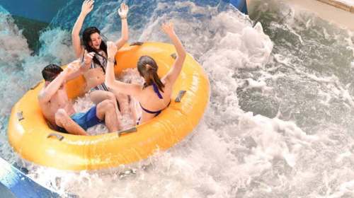 Three people in a yellow inflatable raft enjoy a water slide, splashing in the waves with hands raised in excitement.