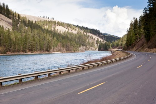A winding road alongside a river, surrounded by lush green trees and rolling hills under a partly cloudy sky.