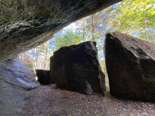 A view from inside a rocky cave, with large boulders and sunlight filtering through trees above.