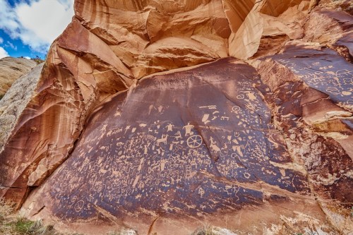 Ancient petroglyphs etched into a rocky cliff, showcasing various symbols and figures against a reddish-brown background.
