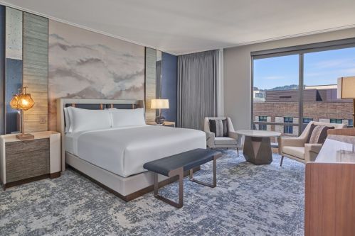 A modern hotel room featuring a king bed, seating area, and large window with natural light. Stylish decor and furnishings.