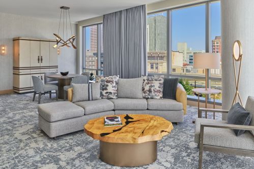 A modern hotel room featuring a gray sectional sofa, wooden coffee table, and large windows with city views.