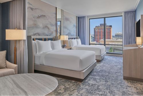 A modern hotel room featuring two beds, a seating area, and large windows with a city view.