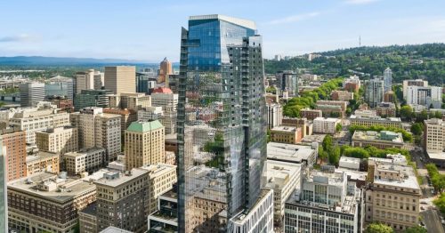 Aerial view of a modern skyscraper in a city, surrounded by greenery and urban buildings under a clear blue sky.