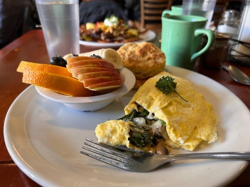A plate with a vegetable omelet, fruit bowl with apple and orange slices, a biscuit, and a green mug.