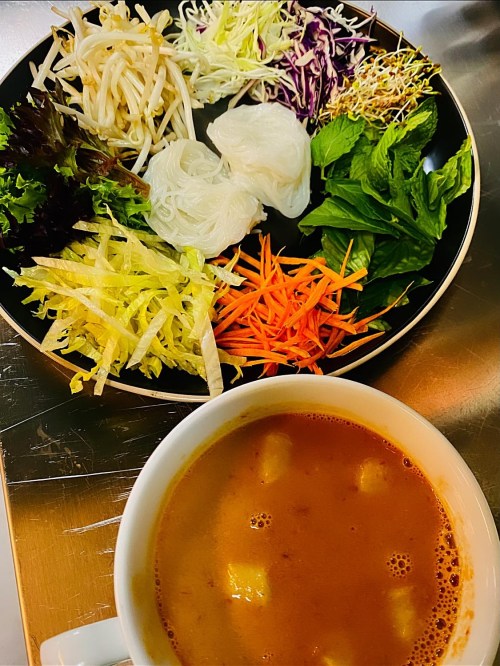 A bowl of soup with tofu, surrounded by a plate of fresh vegetables and herbs, including lettuce, carrots, and bean sprouts.