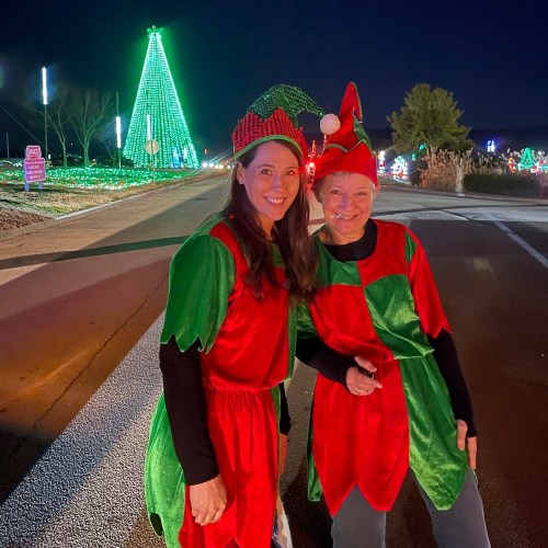 Two women dressed as elves in festive costumes pose together in front of a brightly lit Christmas tree at night.