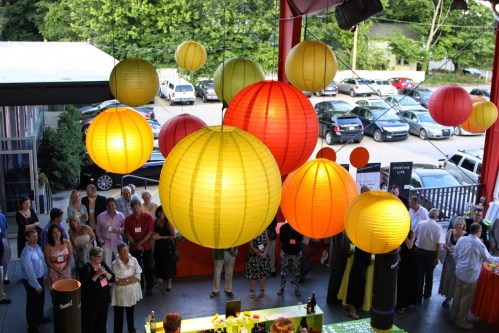Colorful lanterns hang above a crowd at an outdoor event, with cars parked in the background and greenery surrounding the area.