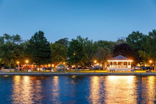 A serene lakeside scene at dusk, featuring a gazebo, trees, and warm lights reflecting on the water.