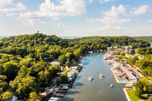 Aerial view of a serene river lined with trees, homes, and boats, with a hill and blue sky in the background.