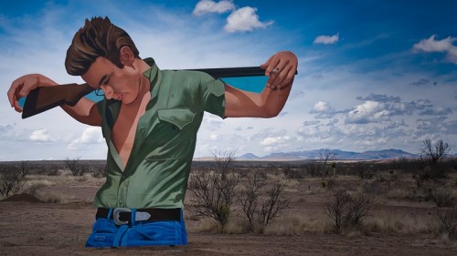 A large mural of a man in a green shirt holding a rifle, set against a desert landscape and cloudy sky.