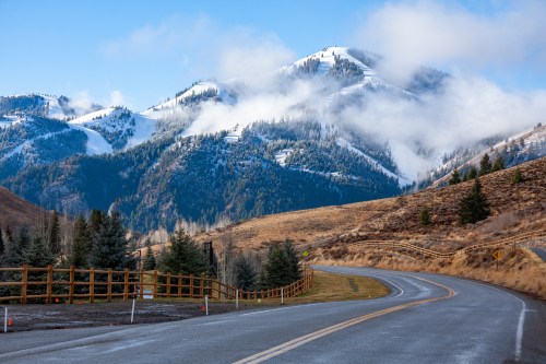 A winding road leads through a mountainous landscape with snow-capped peaks and clouds under a clear blue sky.