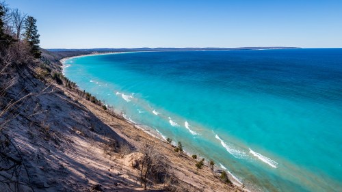 A scenic view of a turquoise lake meeting a sandy shoreline, surrounded by trees under a clear blue sky.