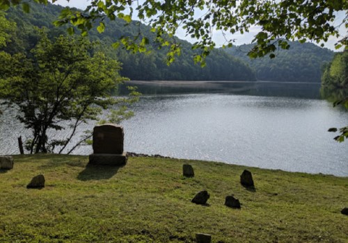 A serene lakeside scene with a gravestone and scattered stones, surrounded by trees and mountains in the background.