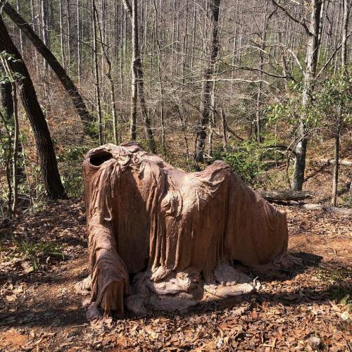 A draped sculpture resembling a figure, partially covered in fabric, set in a wooded area with trees and fallen leaves.