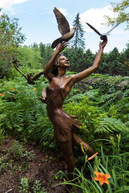 A bronze sculpture of a joyful woman releasing birds, surrounded by lush greenery and vibrant flowers.