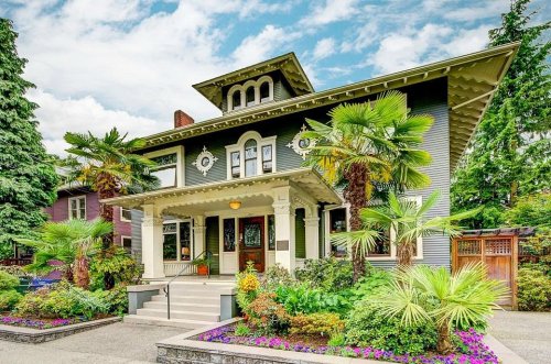 A large, elegant house with a porch, surrounded by lush greenery and colorful flowers under a partly cloudy sky.
