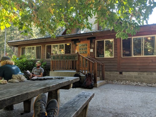A rustic bakery with large windows, surrounded by trees, and a picnic table in the foreground. Two people are seated nearby.