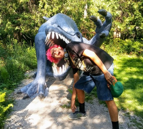 A person with pink hair poses playfully as if being eaten by a large, cartoonish monster sculpture in a green park.