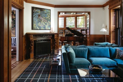 A cozy living room with a teal sofa, wooden accents, a grand piano, and a large window letting in natural light.