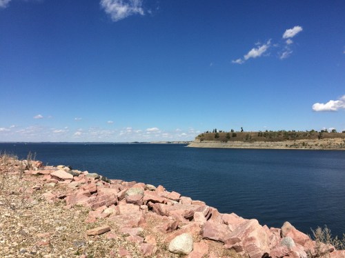 A serene lake scene with rocky shoreline, clear blue water, and a bright sky dotted with clouds.