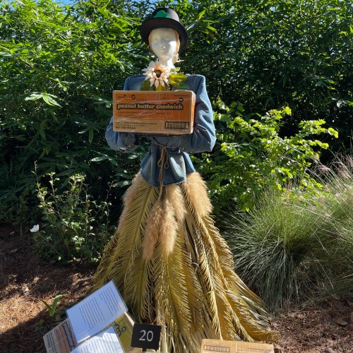 A mannequin dressed in a blue jacket and straw skirt holds a box labeled "peanut butter sandwich" with a flower.