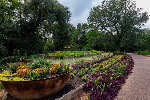 A lush garden with colorful flower beds and greenery, bordered by stone paths and large planters.